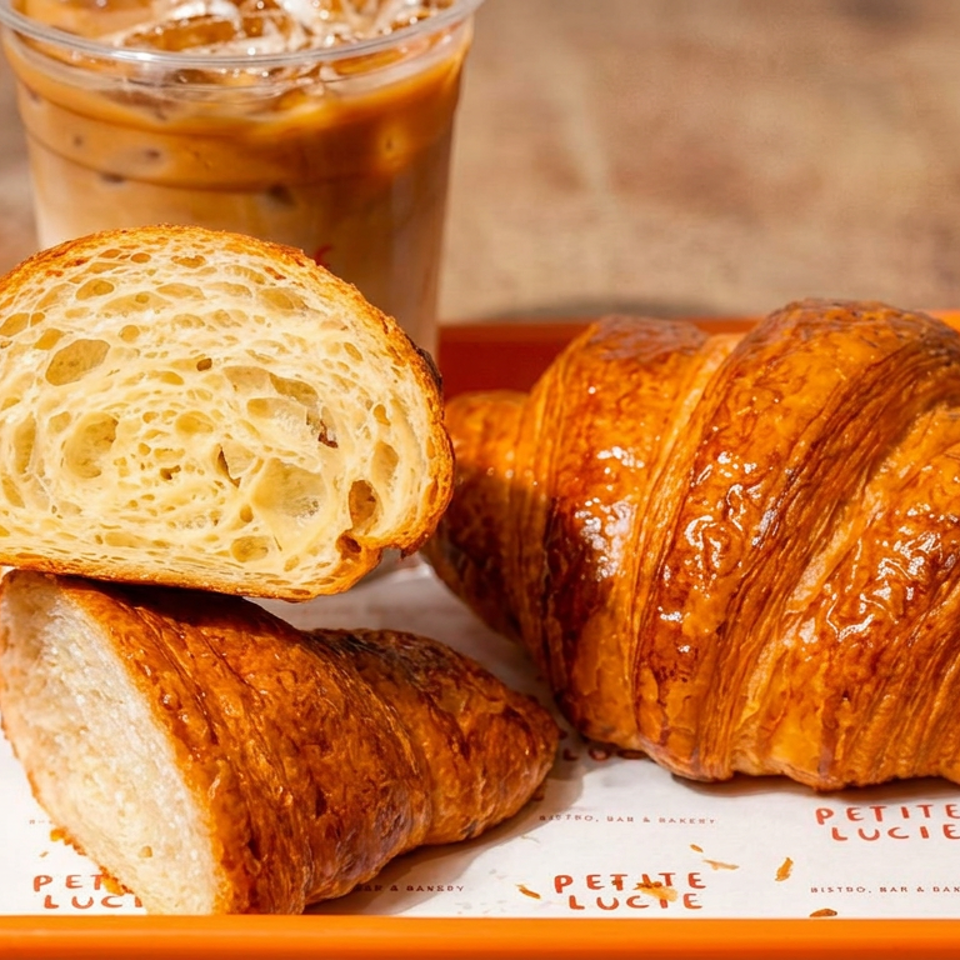 Golden croissants and sliced crusty bread served on a branded orange tray, with an iced coffee in the background.